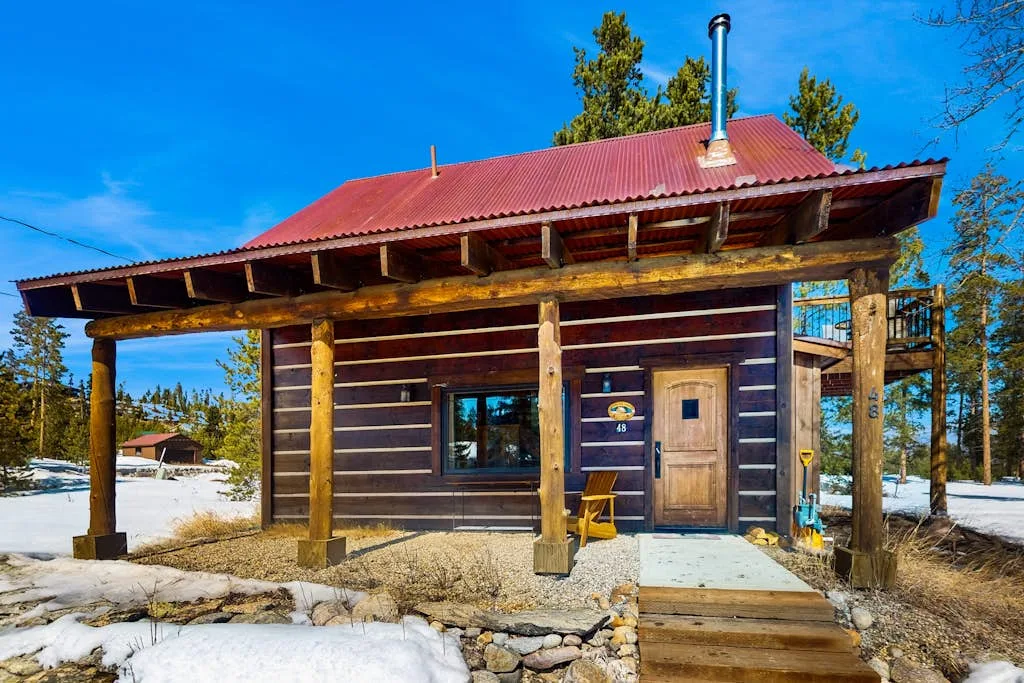 A rustic wooden cabin stands beneath a red metal roof in a snowy landscape.
