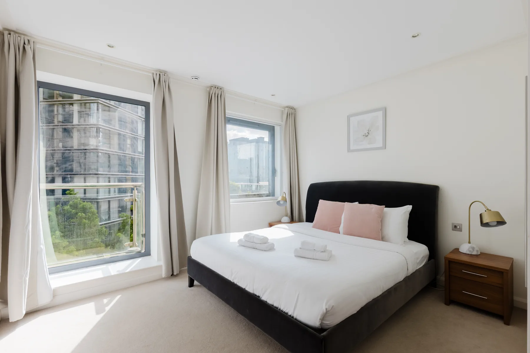 A bright bedroom with a large bed, pale curtains, and windows overlooking nearby buildings.