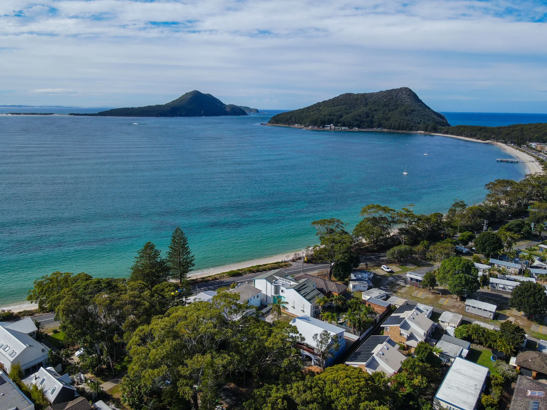Aerial coastal view of a turquoise bay, wooded headlands, and homes along the shore.