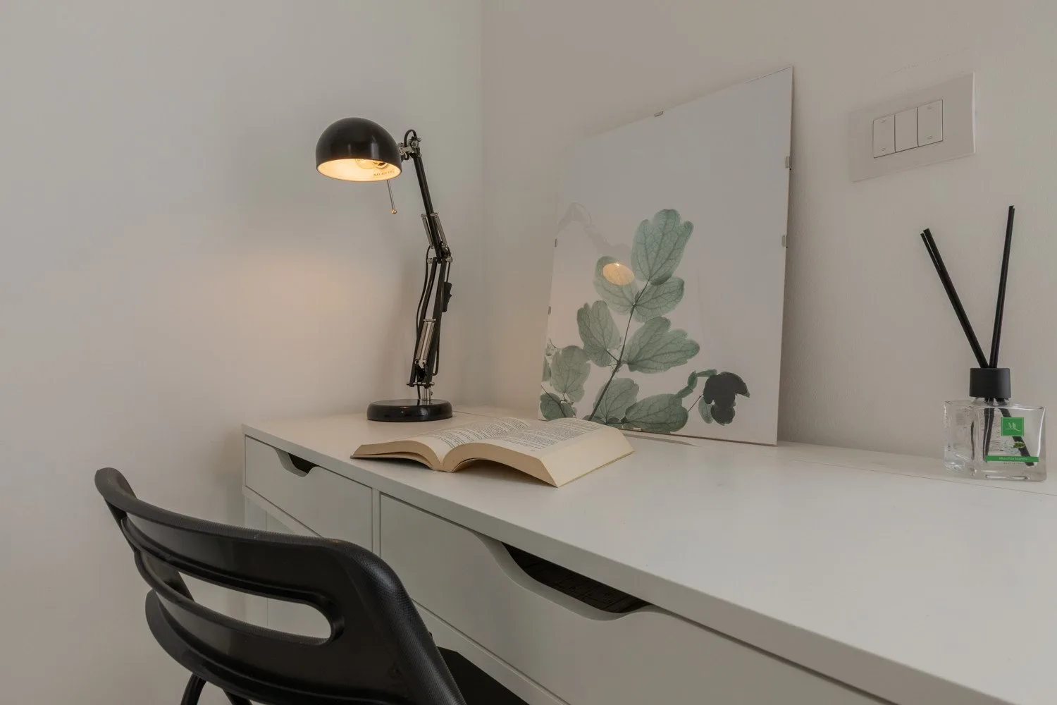 A minimalist desk with an open book, a lamp, and botanical artwork.