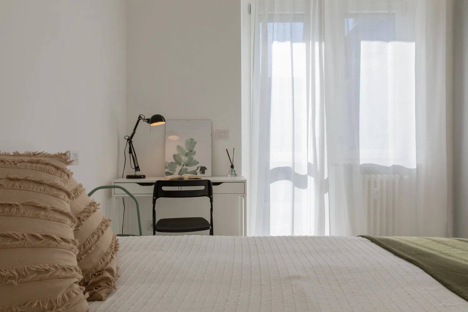 A bedroom with a bed, desk, and sheer curtains over the window.