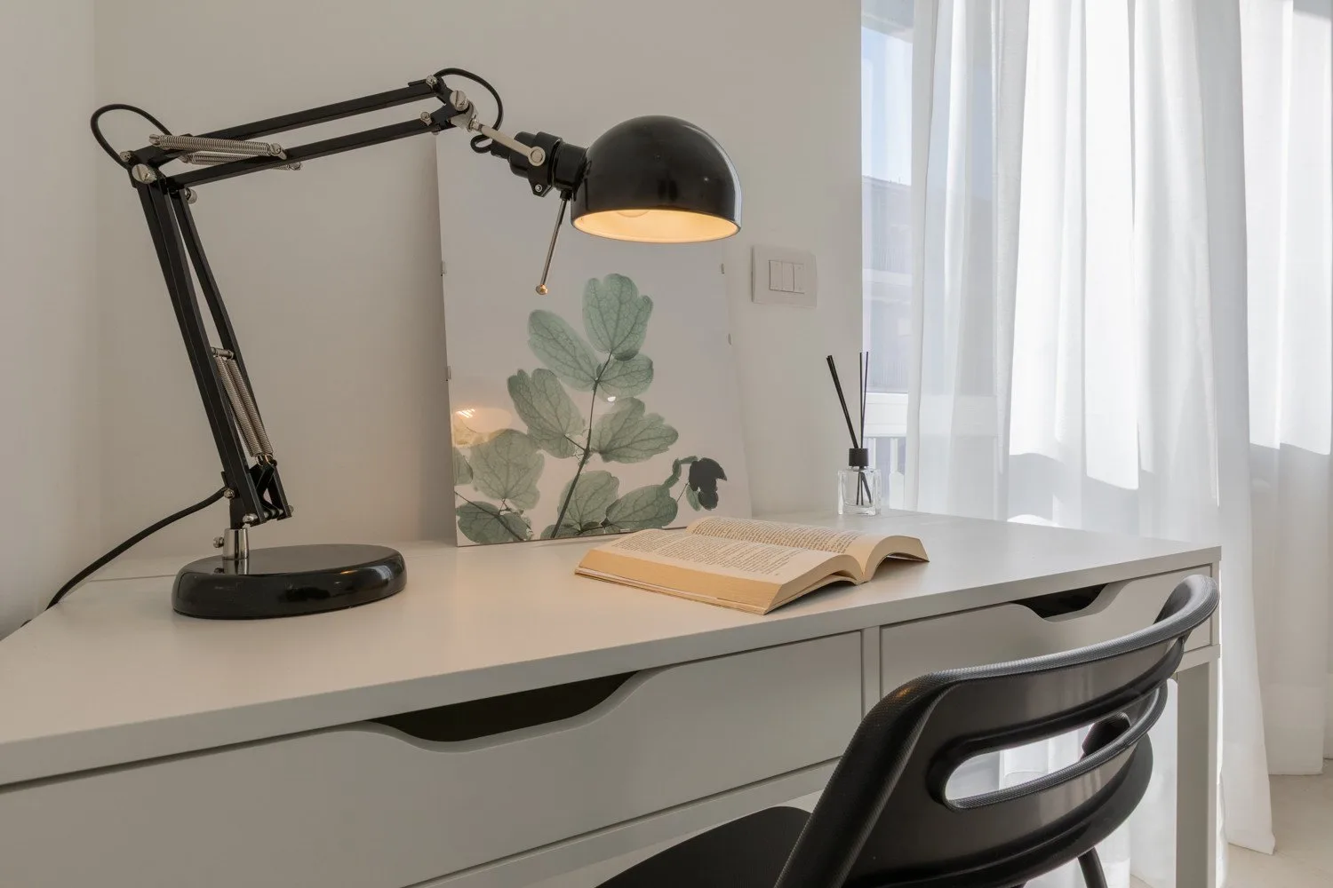 A desk workspace with an open book and an adjustable lamp beside a bright window.
