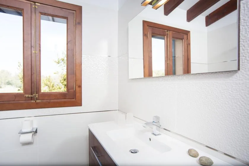A bathroom vanity with a sink sits beneath a large mirror beside a wooden-framed window.