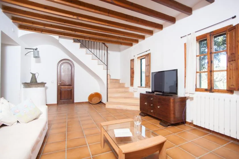 A bright living room with terracotta floor tiles, wooden ceiling beams, and a television.