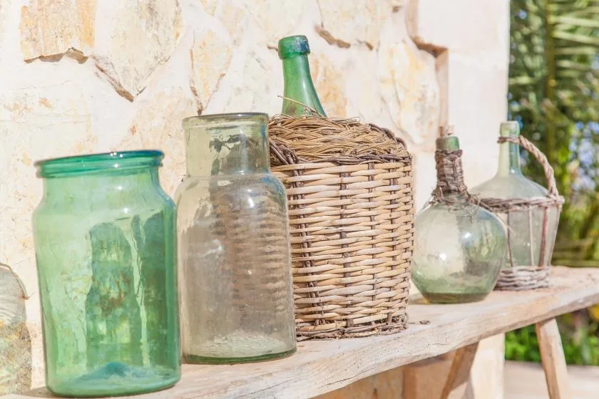 Glass bottles and jars sit on a wooden shelf against a stone wall.