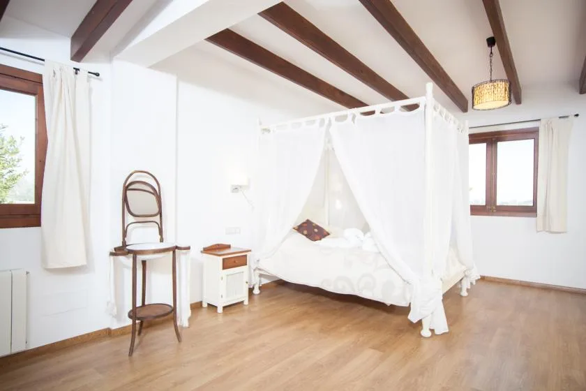 A bright bedroom with a canopy bed, wooden ceiling beams, and windows.