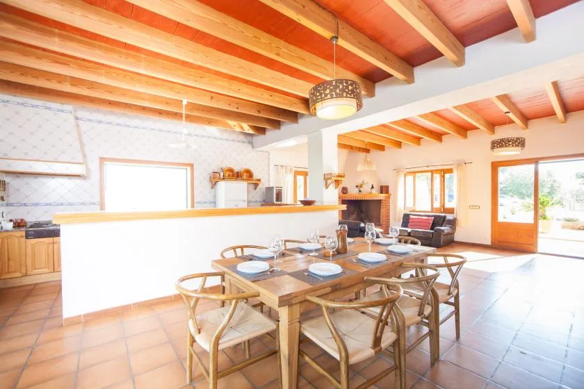 A bright open-plan dining and living area with a long wooden table beneath exposed ceiling beams.