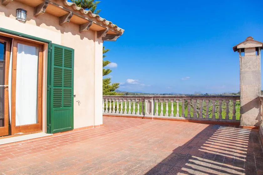 A sunlit terrace with tiled flooring, a green shutter, and a balustrade.