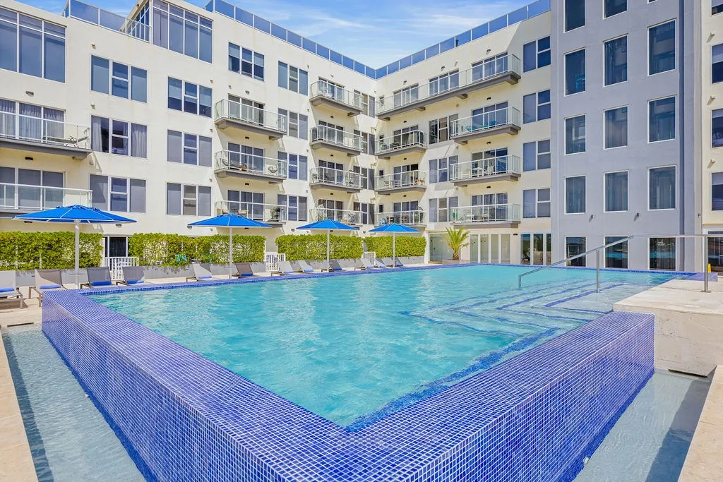 Modern apartment complex featuring a striking glass-walled pool in an inner courtyard. The pool area is surrounded by a well-maintained landscaped hedge, vibrant blue umbrellas, and loungers—ideal for relaxation under sunny skies.