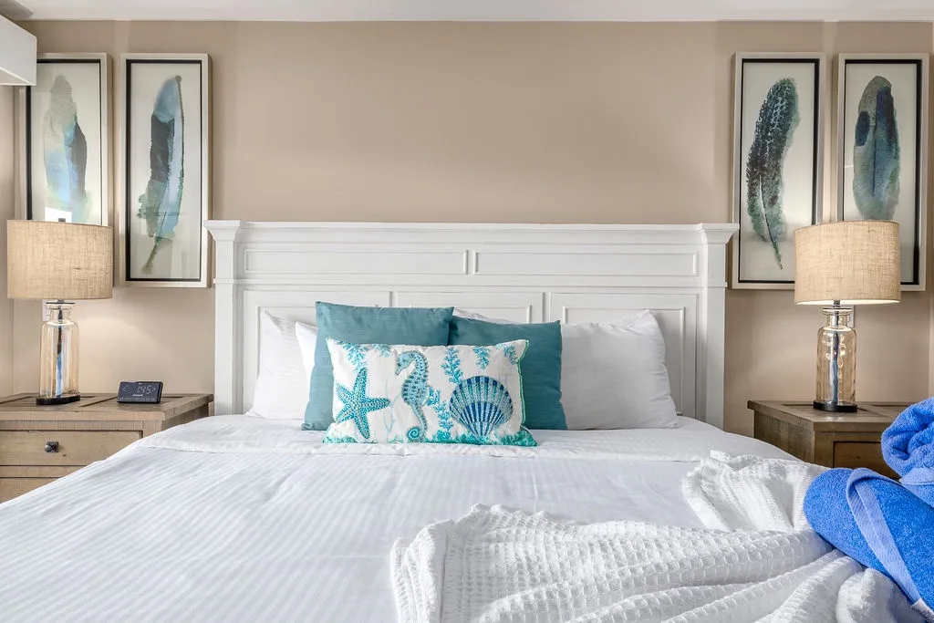 Sink into crisp white linens and coastal charm, where soft lighting and serene coastal art invite your restful escape.