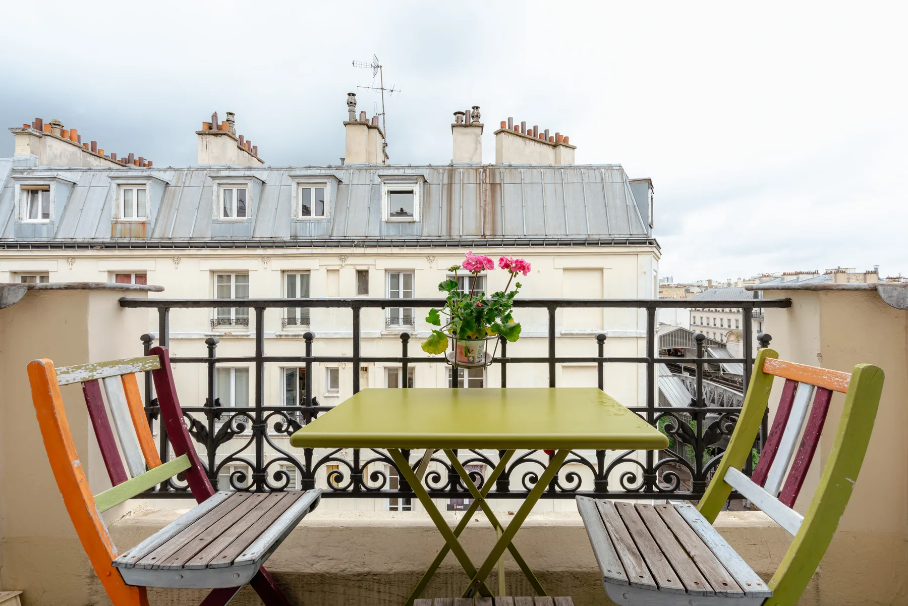 A balcony dining area with a small table, colorful seating, and potted flowers.