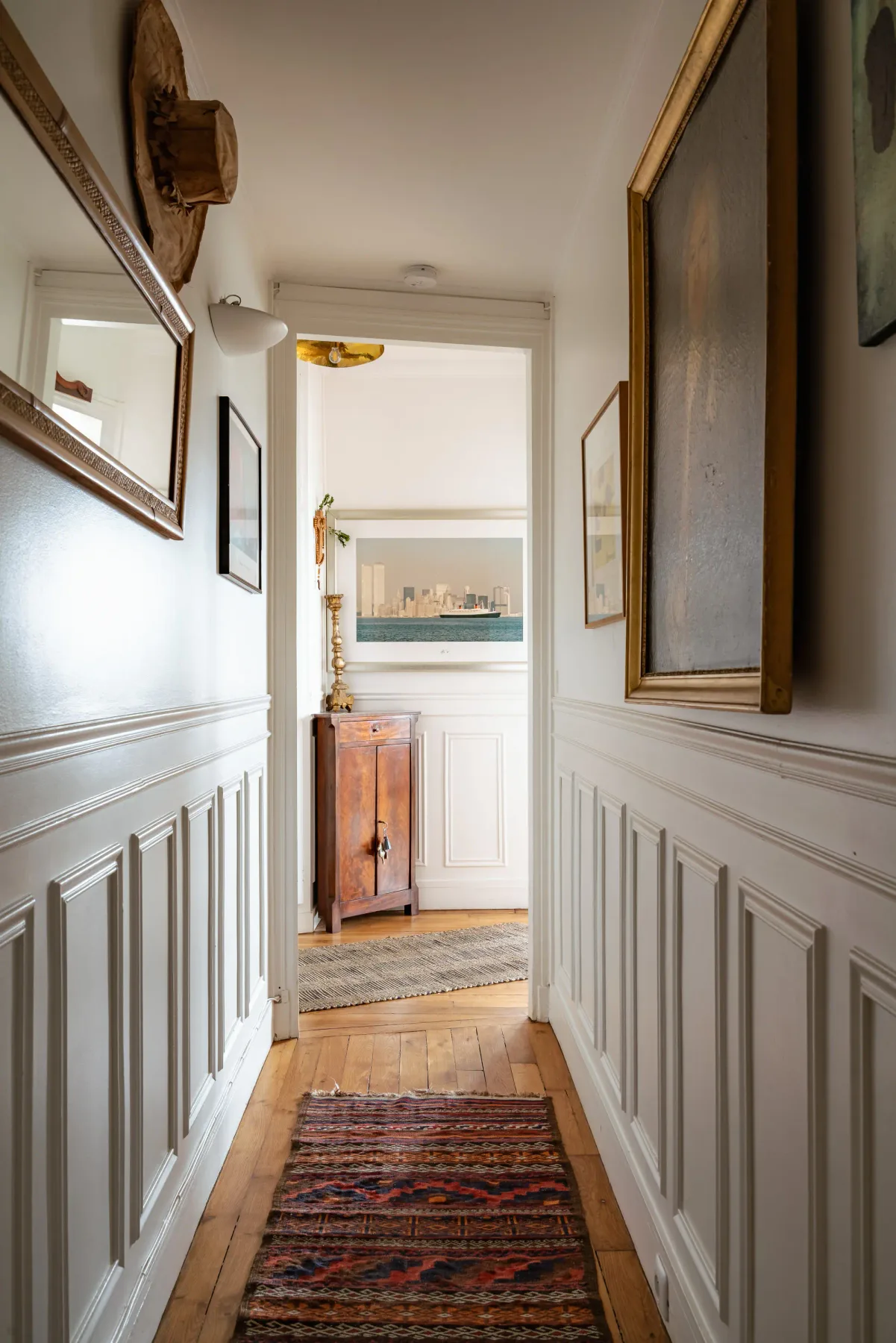 A bright hallway with white paneled walls, a wooden floor, and framed artwork.