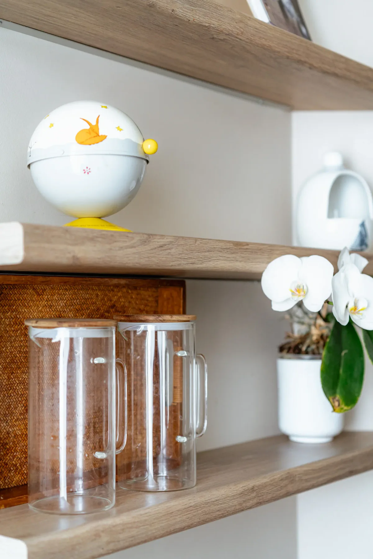 A wooden shelving unit displays clear drink pitchers, a small figurine, and a flowering orchid.
