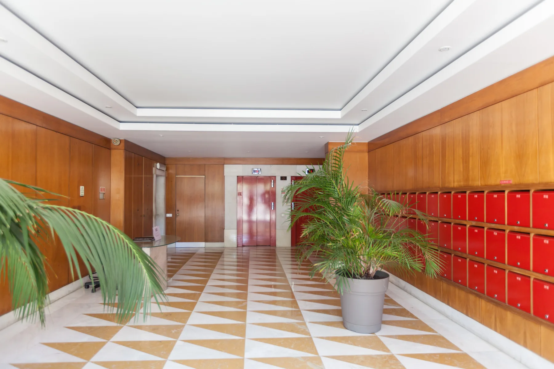 Vestíbulo de un edificio de apartamentos con suelo de baldosas estampadas, plantas y buzones rojos.