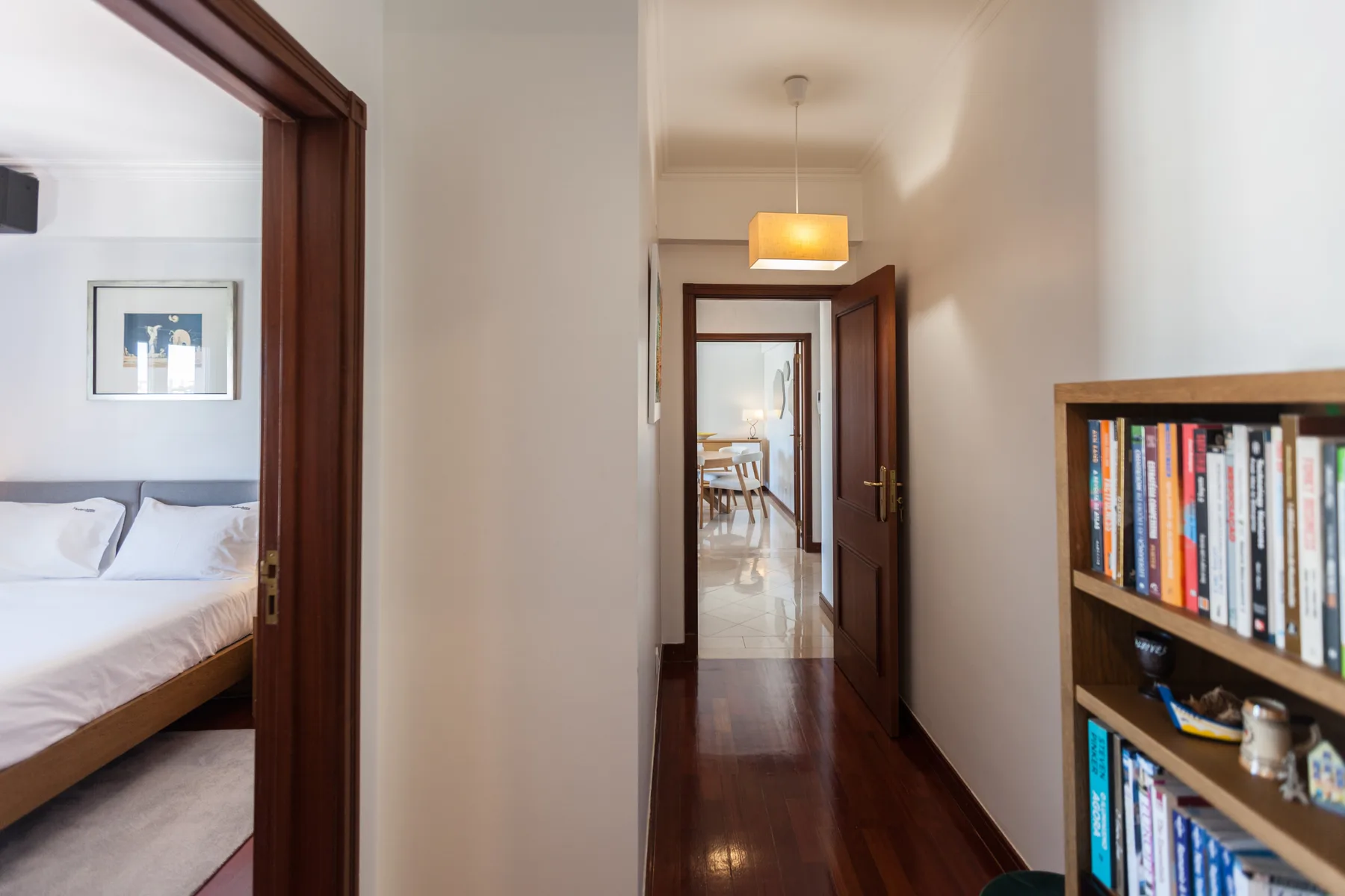 Un pasillo con puertas abiertas, una estantería y un dormitorio visible a un lado.