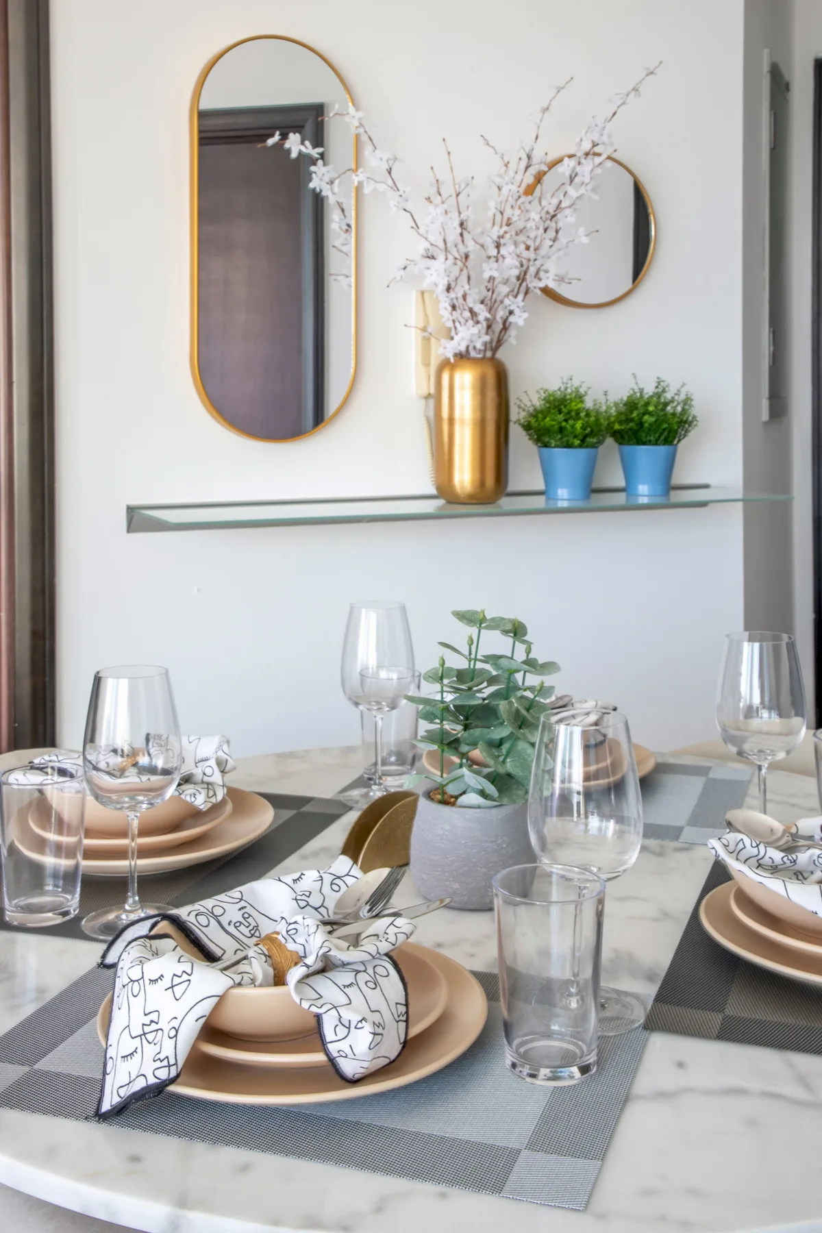 A dining table is set with plates and glasses beneath a wall shelf with plants.