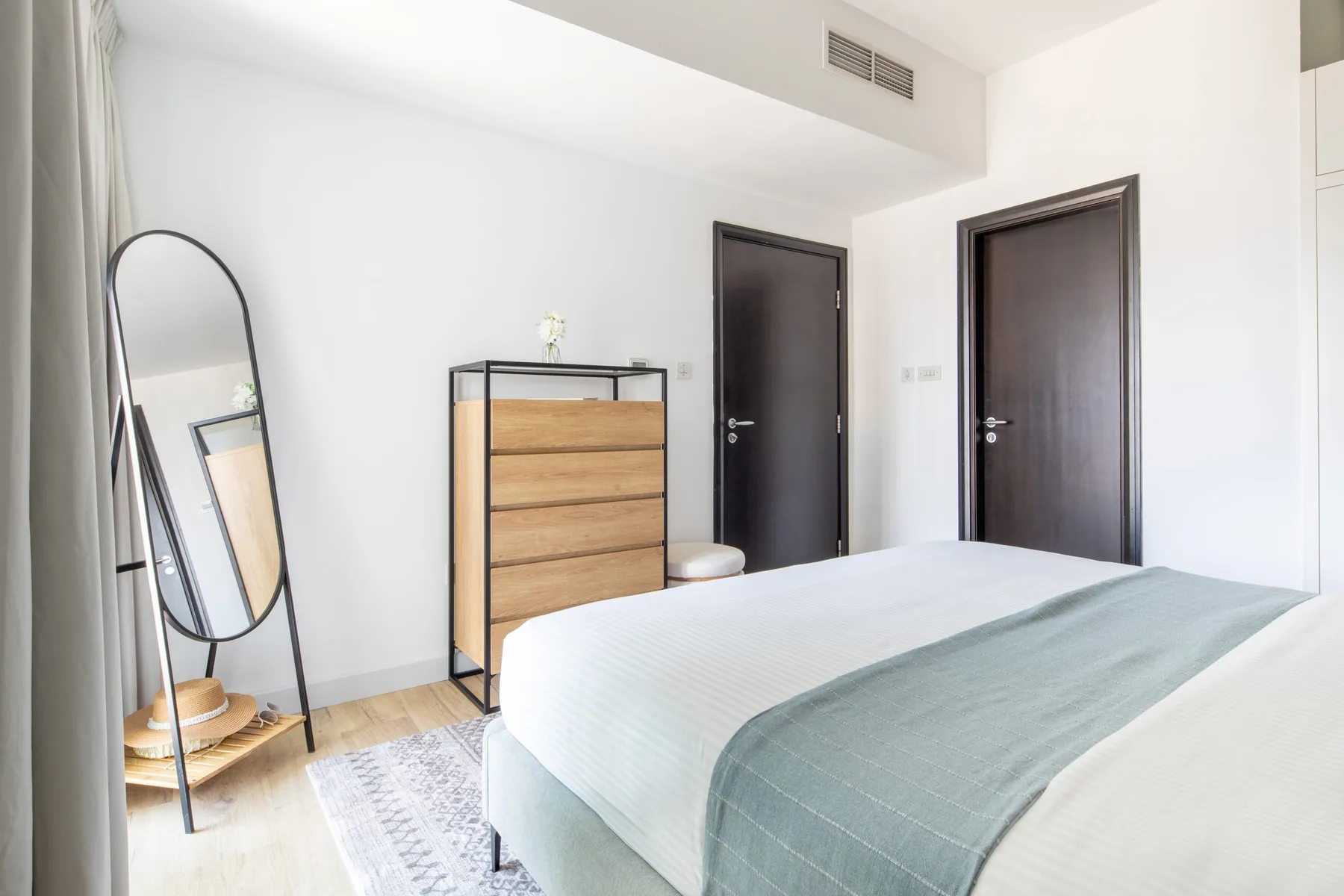 A bright bedroom with a bed, wooden dresser, and two dark doors.