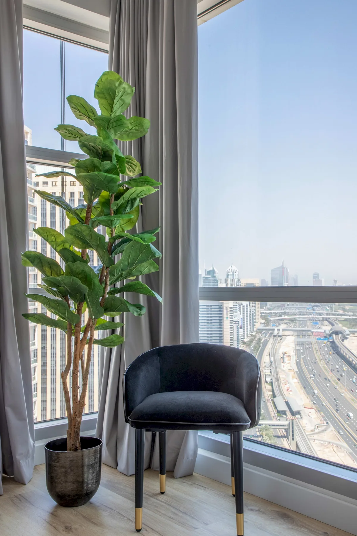 A chair and leafy plant sit beside a floor-to-ceiling window overlooking the city.
