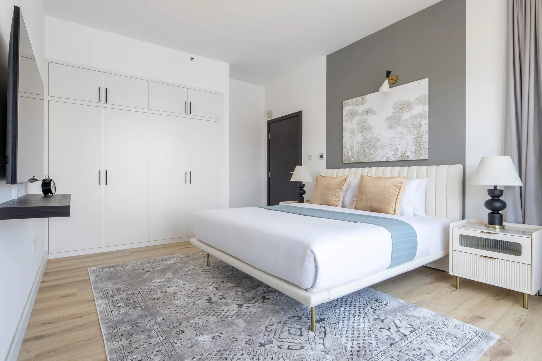 A bright bedroom with a large bed, white wardrobes, and a patterned rug.
