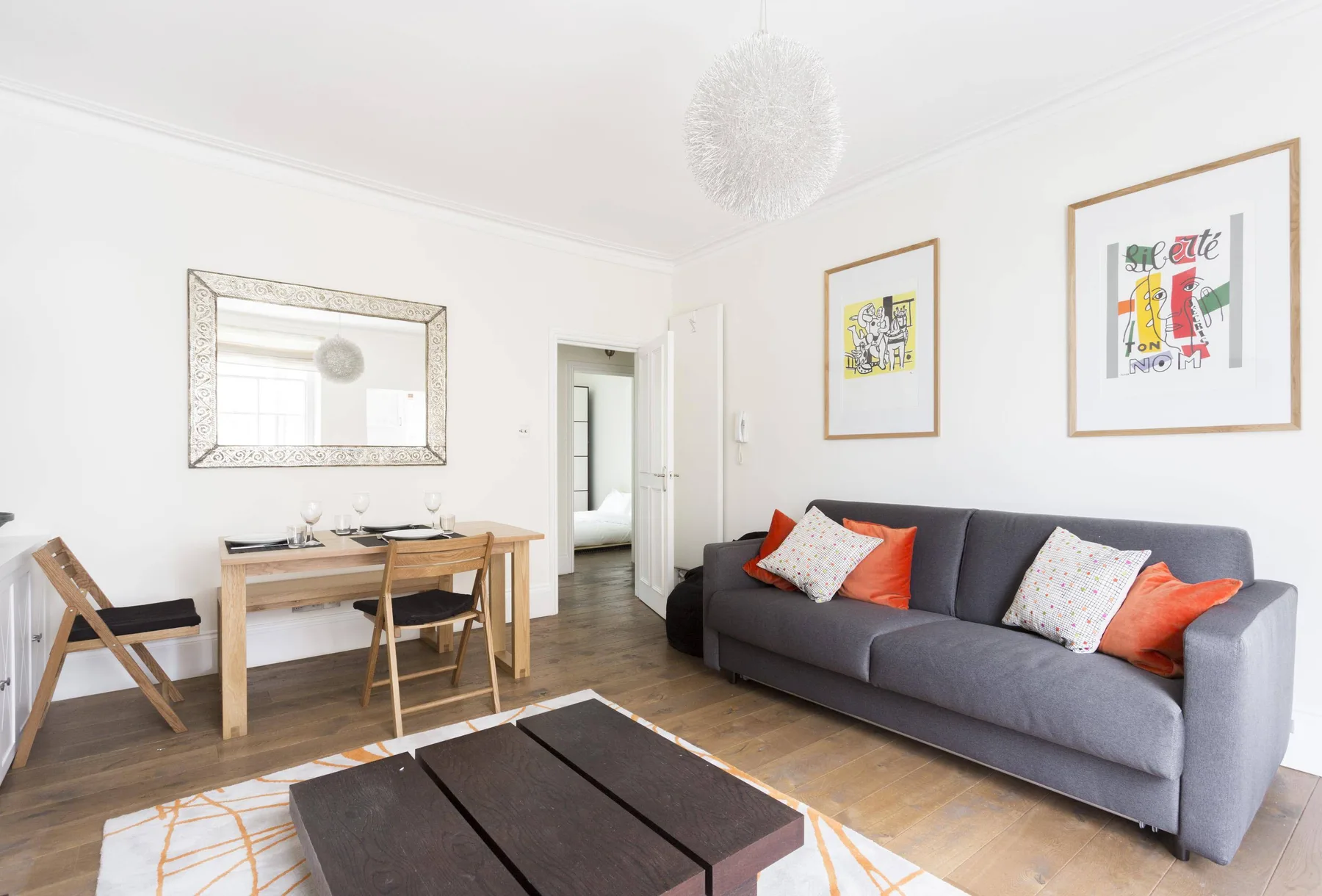 A bright living and dining area with a gray sofa, wooden table, and framed wall art.