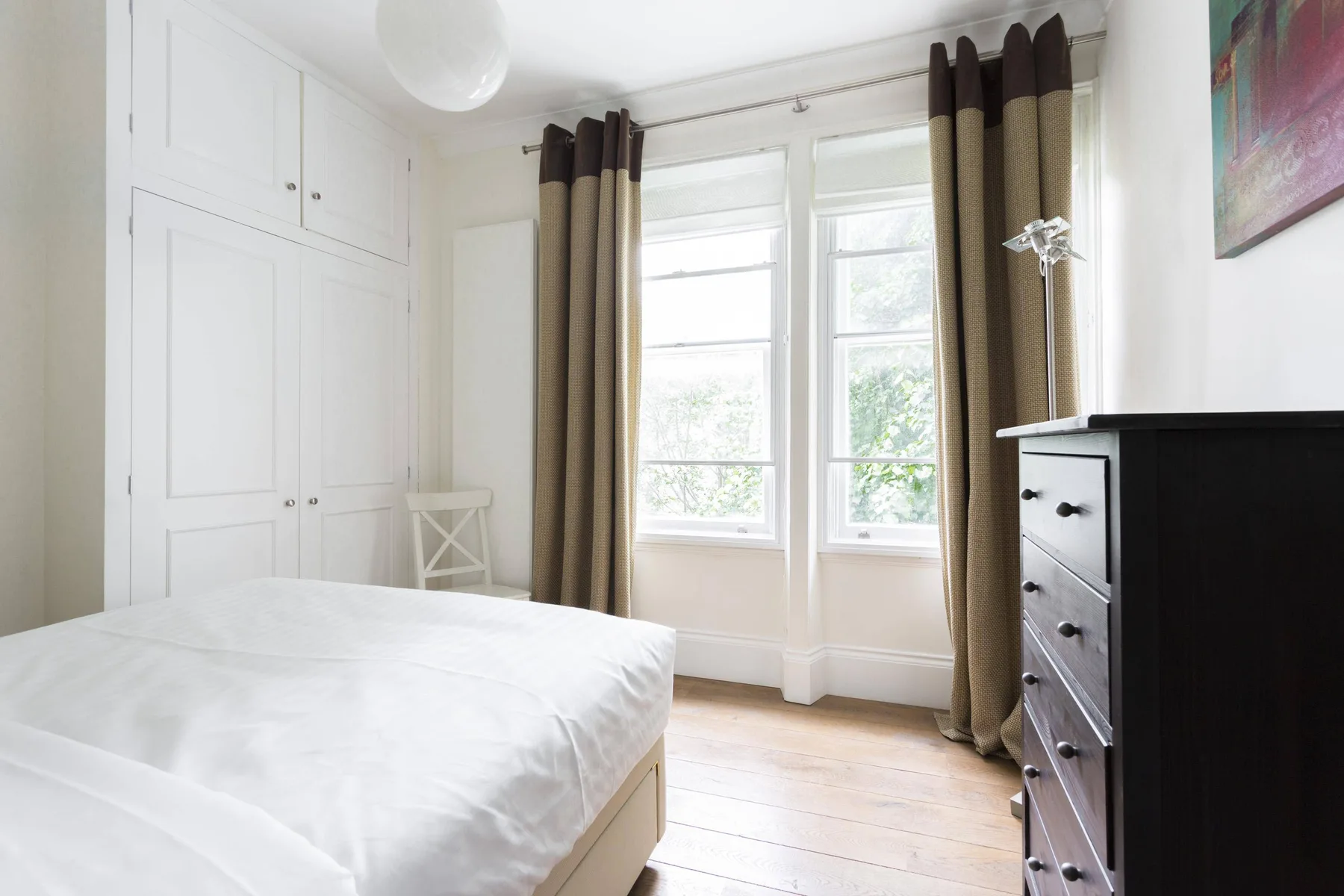 A bright bedroom with a bed, curtained windows, and a dark dresser.