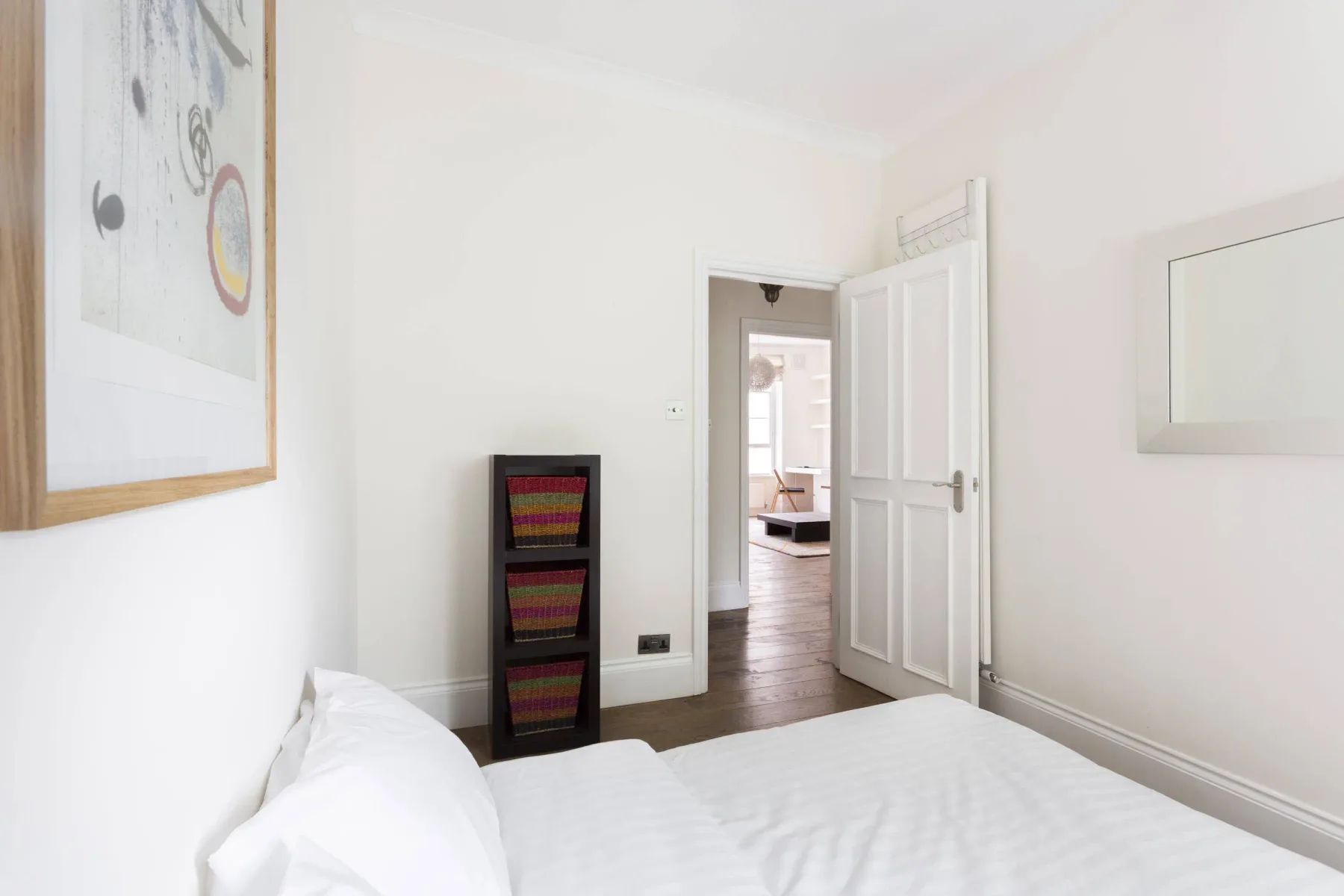 A bright bedroom with white bedding and an open doorway to another room.