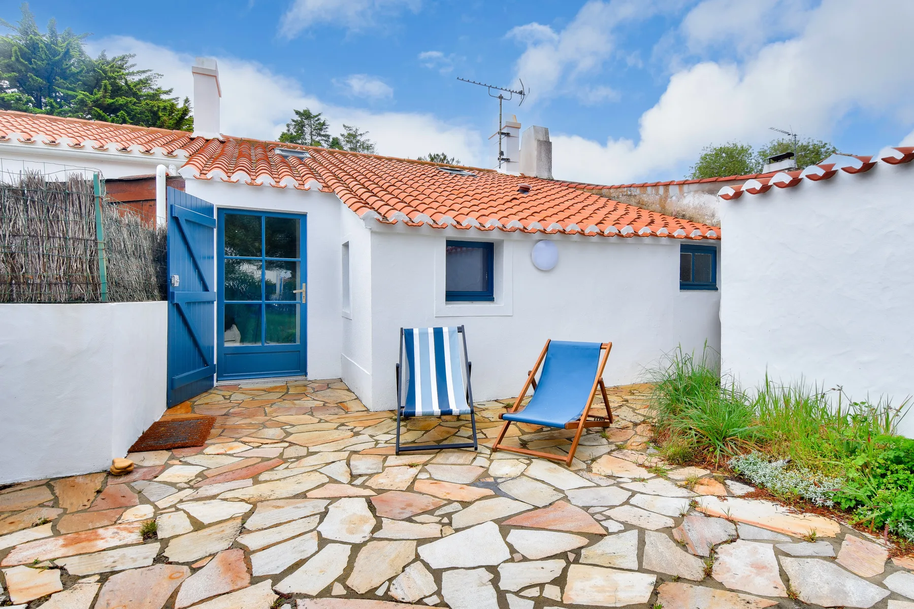 A white house opens onto a stone patio with two striped and blue loungers.
