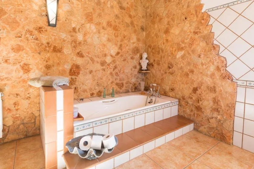 Una bañera empotrada está junto a una pared de textura pétrea en un baño con azulejos.