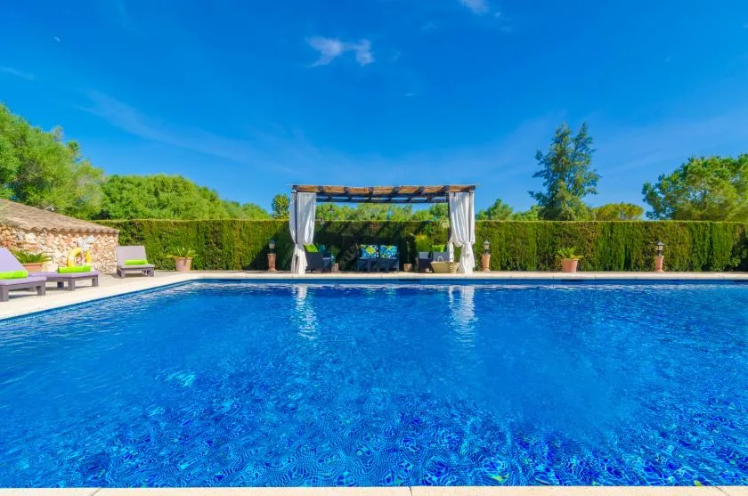 Una piscina junto a una zona de estar con sombra y un jardín ajardinado.