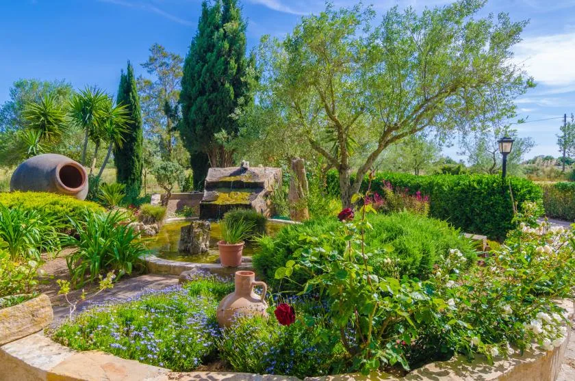 Un jardín frondoso con una pequeña cascada de piedra, plantas con flores y una vasija de barro.