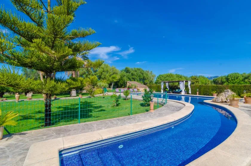 Una piscina exterior curva bordea un césped verde y varios árboles.