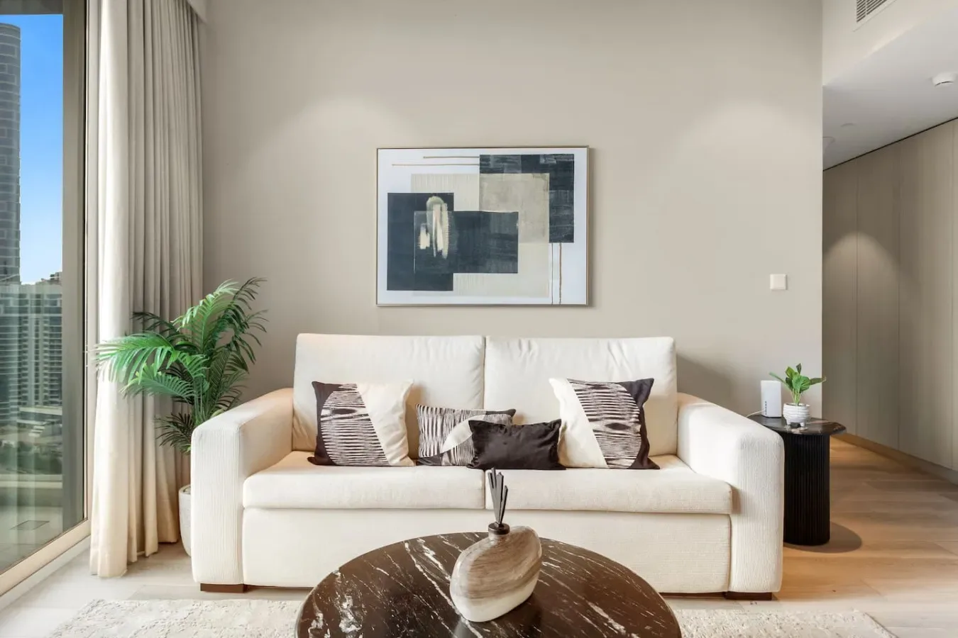 A bright living room with a white sofa, abstract wall art, and a tall window.