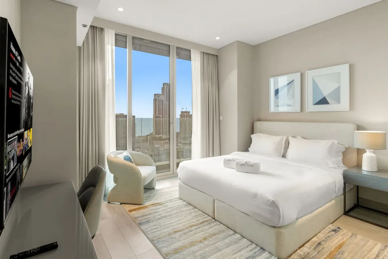 Un dormitorio luminoso con una cama blanca junto a ventanales con vistas a edificios de la ciudad.