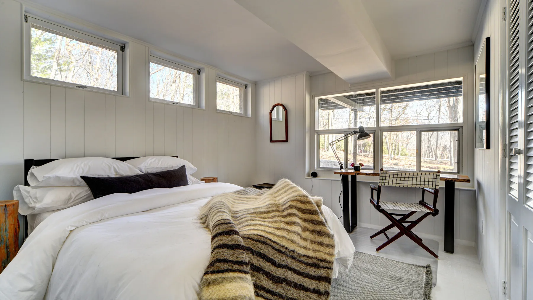 Un dormitorio luminoso con una cama, un escritorio y ventanas grandes.