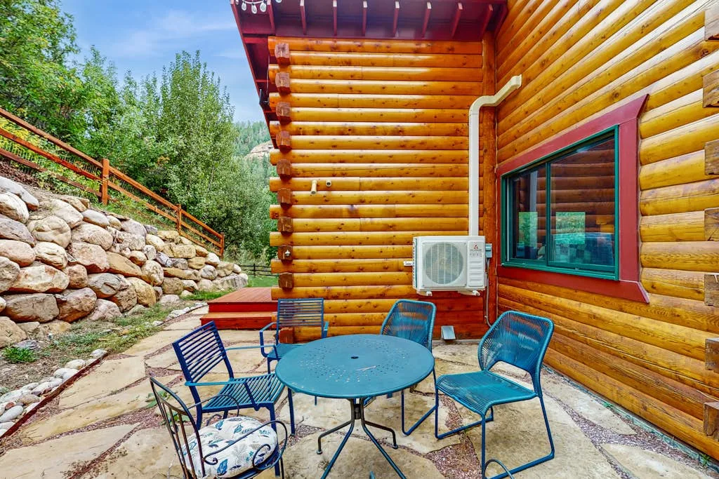 Outdoor seating beside a log cabin, with a stone retaining wall and trees.
