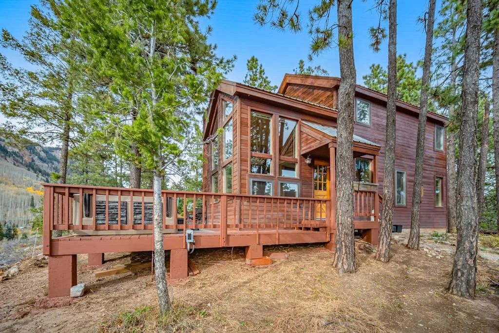 A wood-sided cabin with a raised deck sits among tall pine trees.