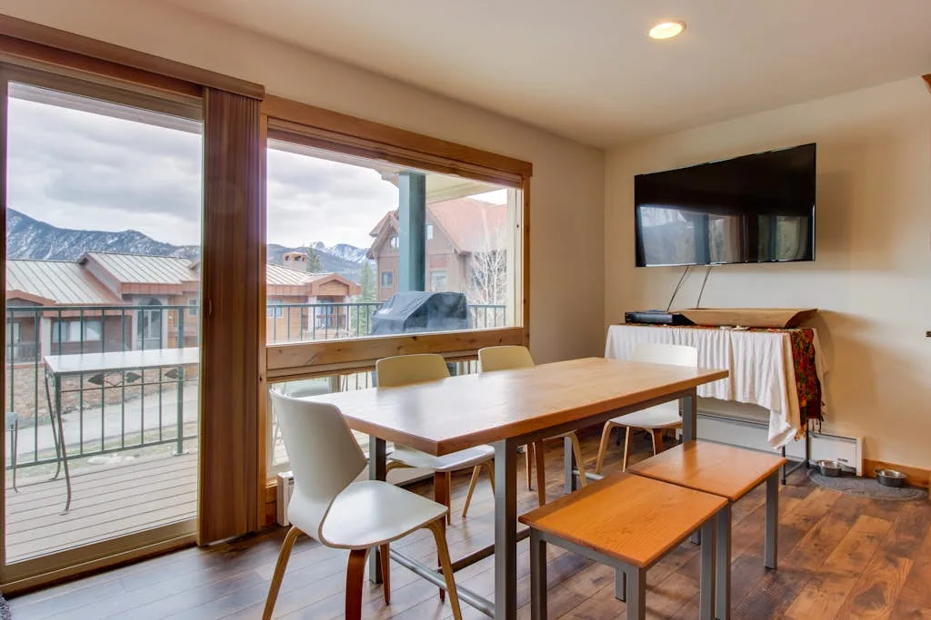 Dining area with a wooden table and chairs beside large windows overlooking nearby buildings.