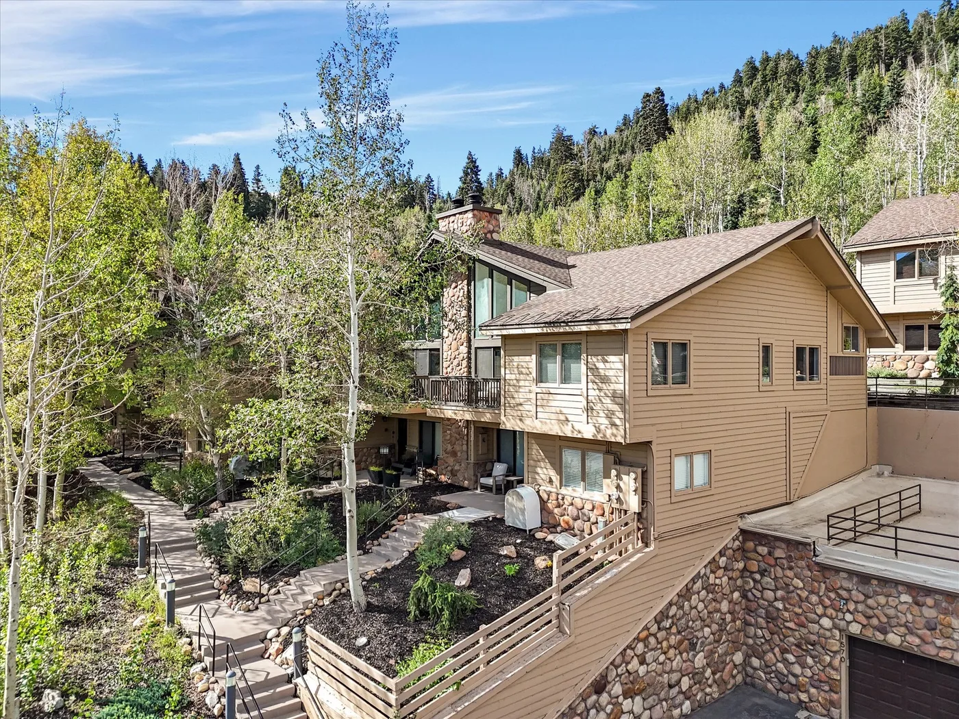 A tan-sided house sits beside a wooded hillside, with a raised wooden walkway.