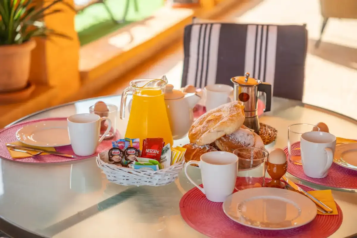 A breakfast table is set with bread, cups, and a coffee pot.