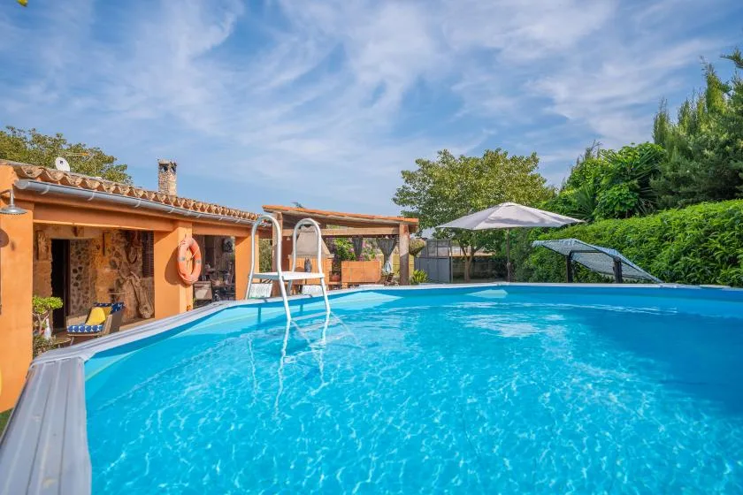 Outdoor swimming pool beside a low building, with trees and umbrellas in the background.