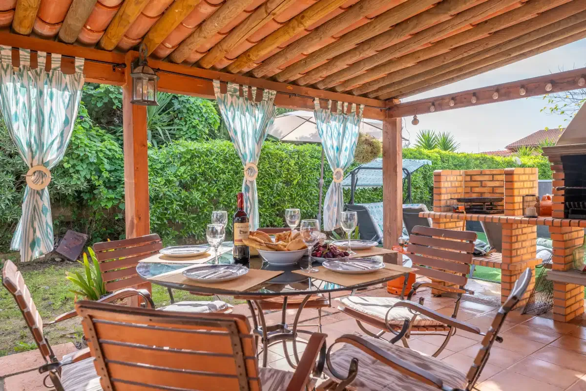 A covered outdoor dining area with a table and chairs beside a brick barbecue.