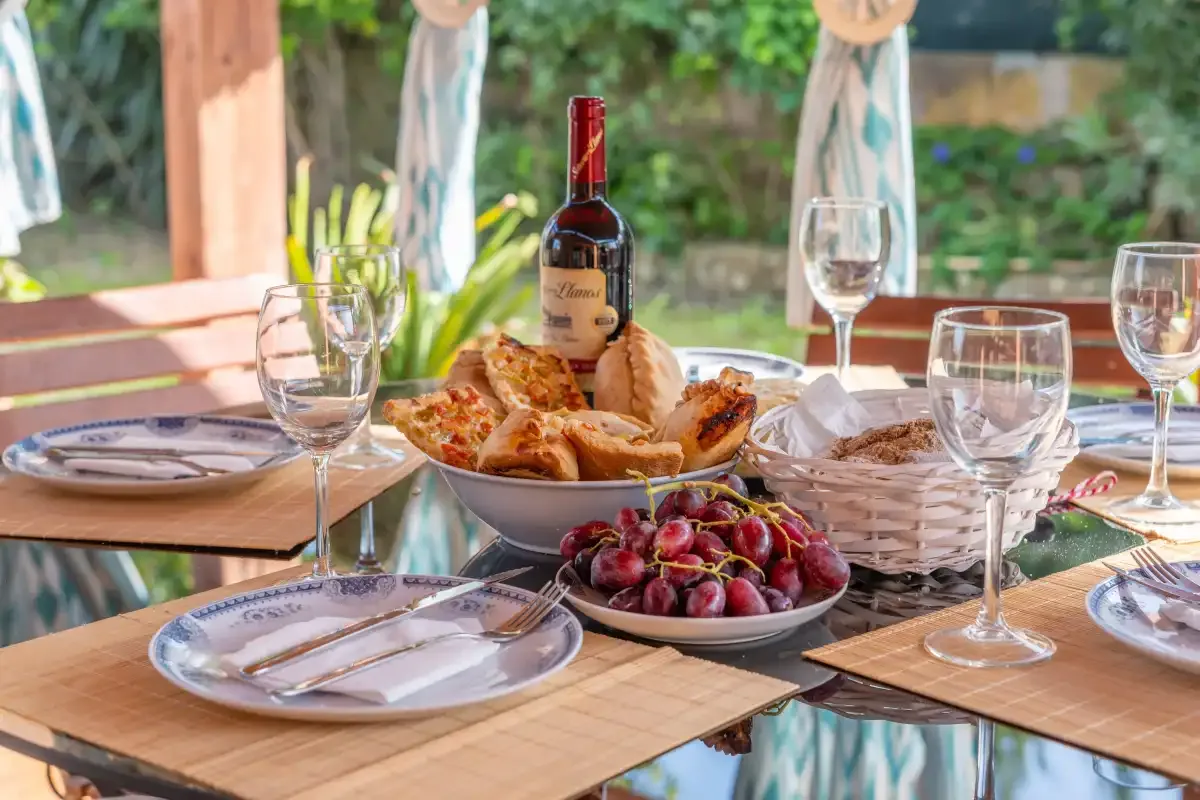 An outdoor dining table is set with food, fruit, and wine glasses.