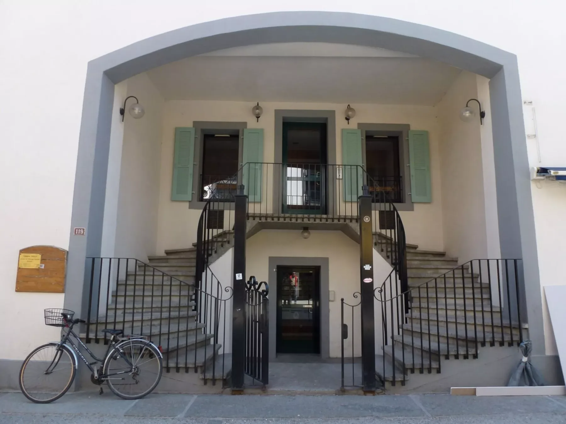 Entrada de un edificio con escaleras a ambos lados, contraventanas verdes y una bicicleta cerca.