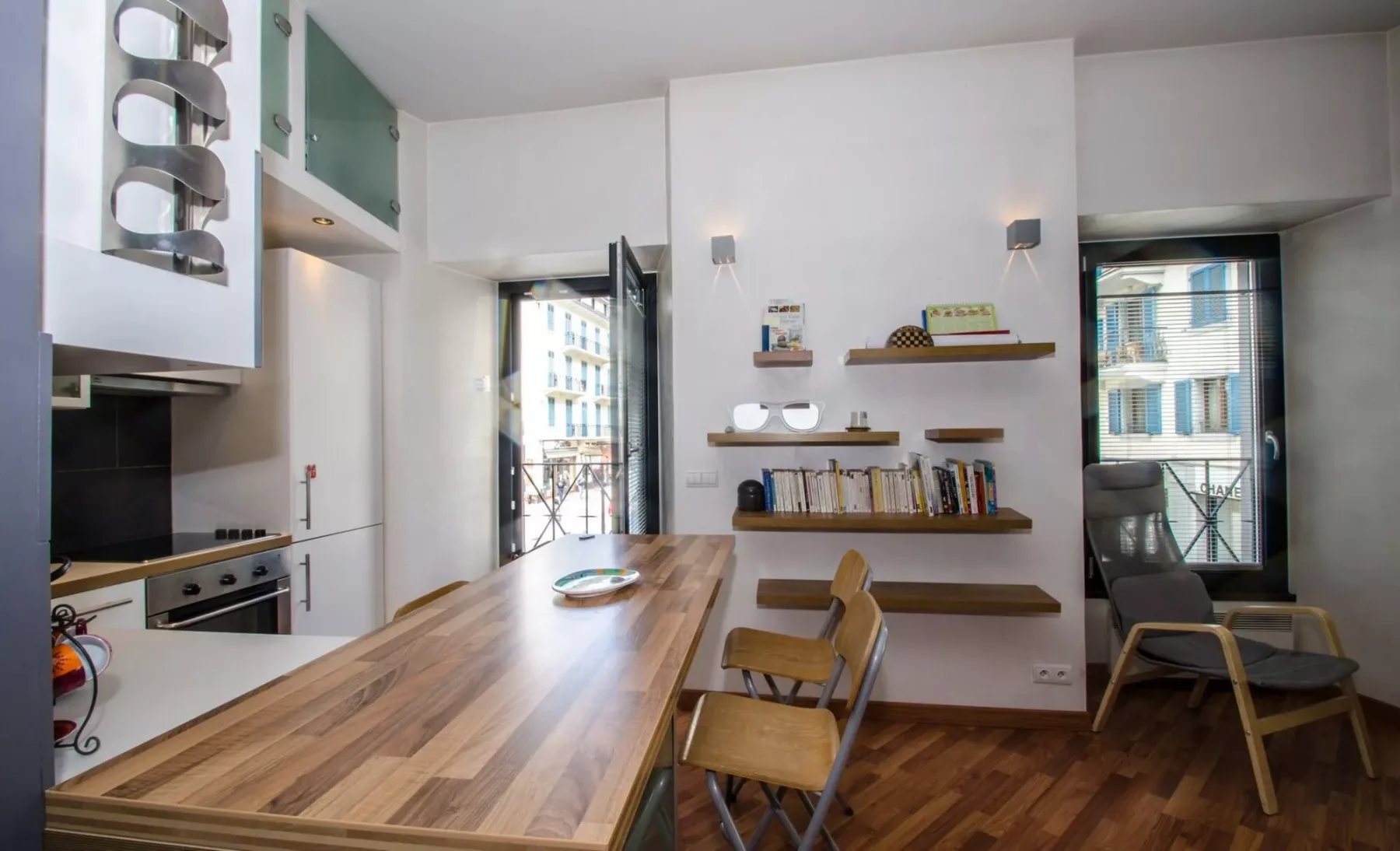 Zona de comedor y cocina con una mesa larga de madera y estantes en la pared.