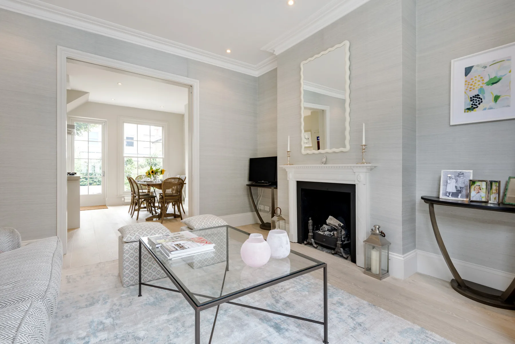 A bright living room with a fireplace, glass coffee table, and dining area beyond.