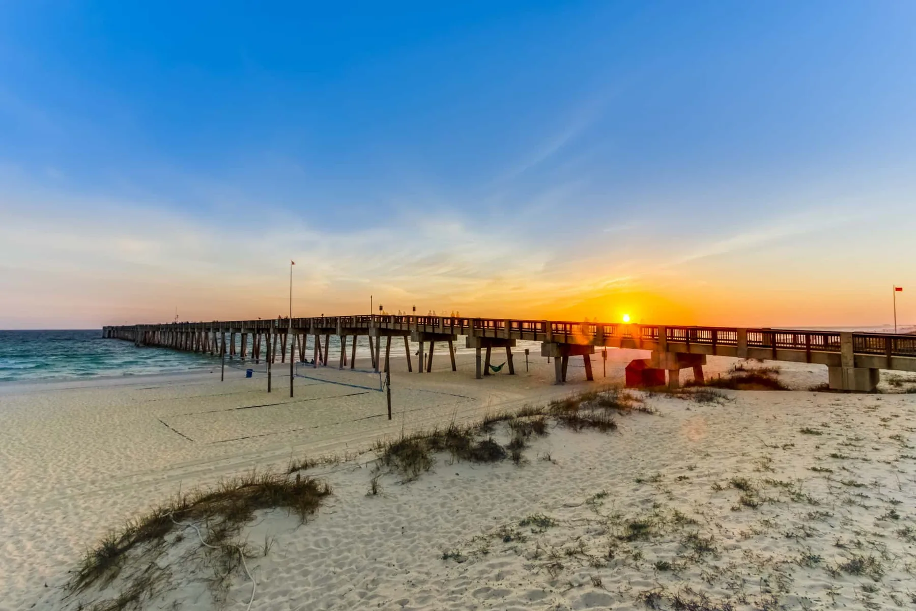 A wooden pier extends over the beach as the sun sets over the ocean.