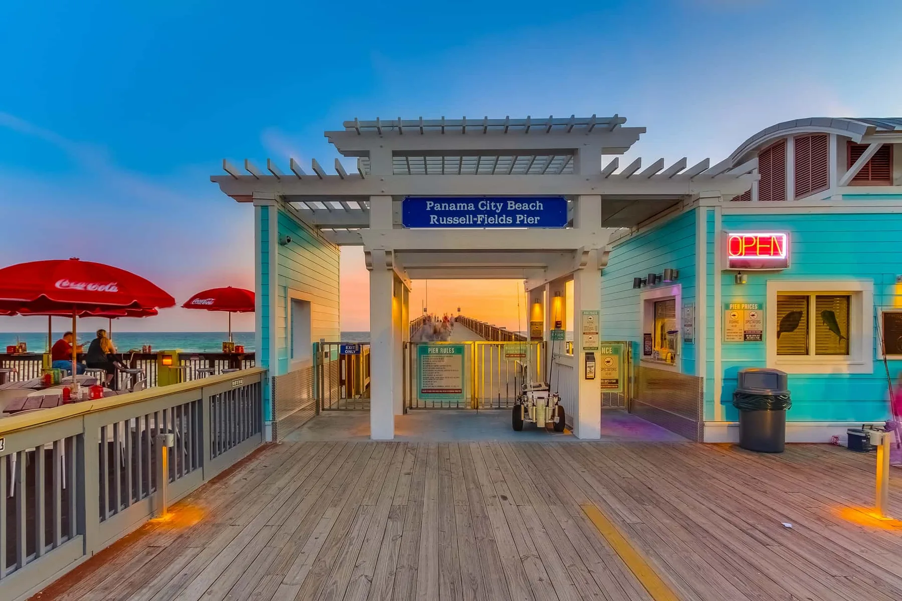 A seaside pier entrance opens onto a wooden boardwalk, with a brightly lit storefront nearby.