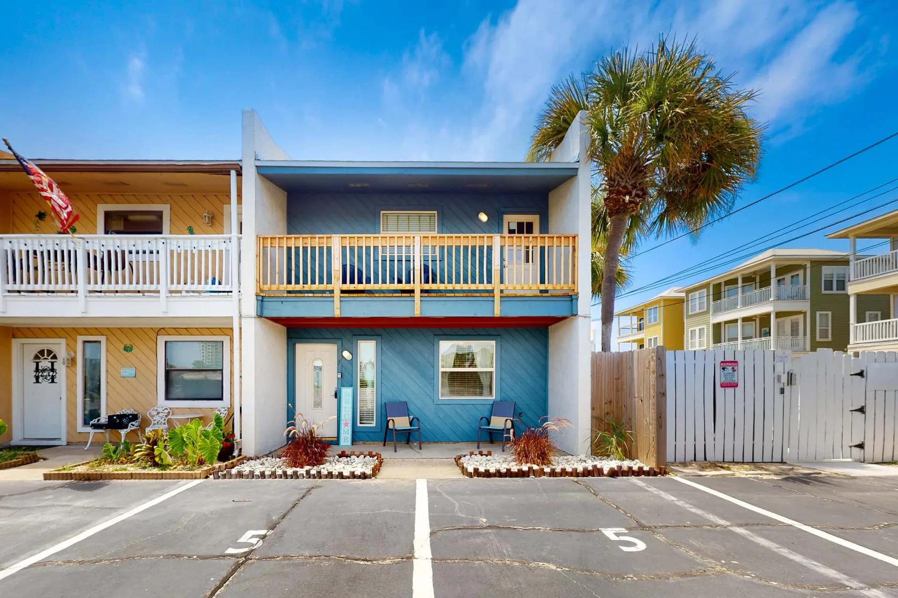 Exterior view of a two-story blue building with balconies and a palm tree.