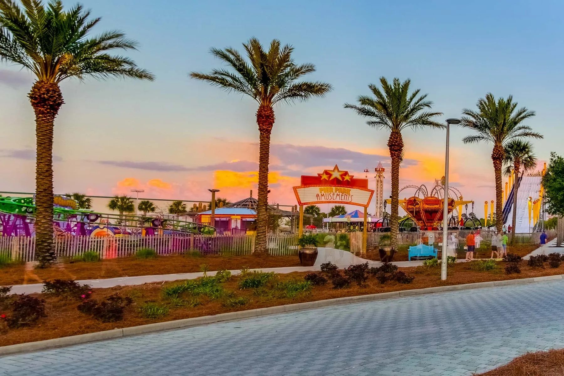 A palm-lined outdoor amusement park entrance beneath a colorful sunset sky.