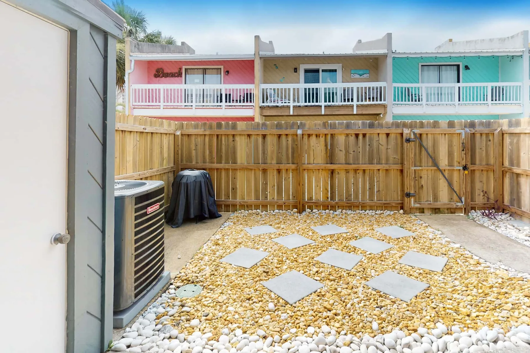 A fenced outdoor gravel patio has stepping stones and neighboring colorful buildings.